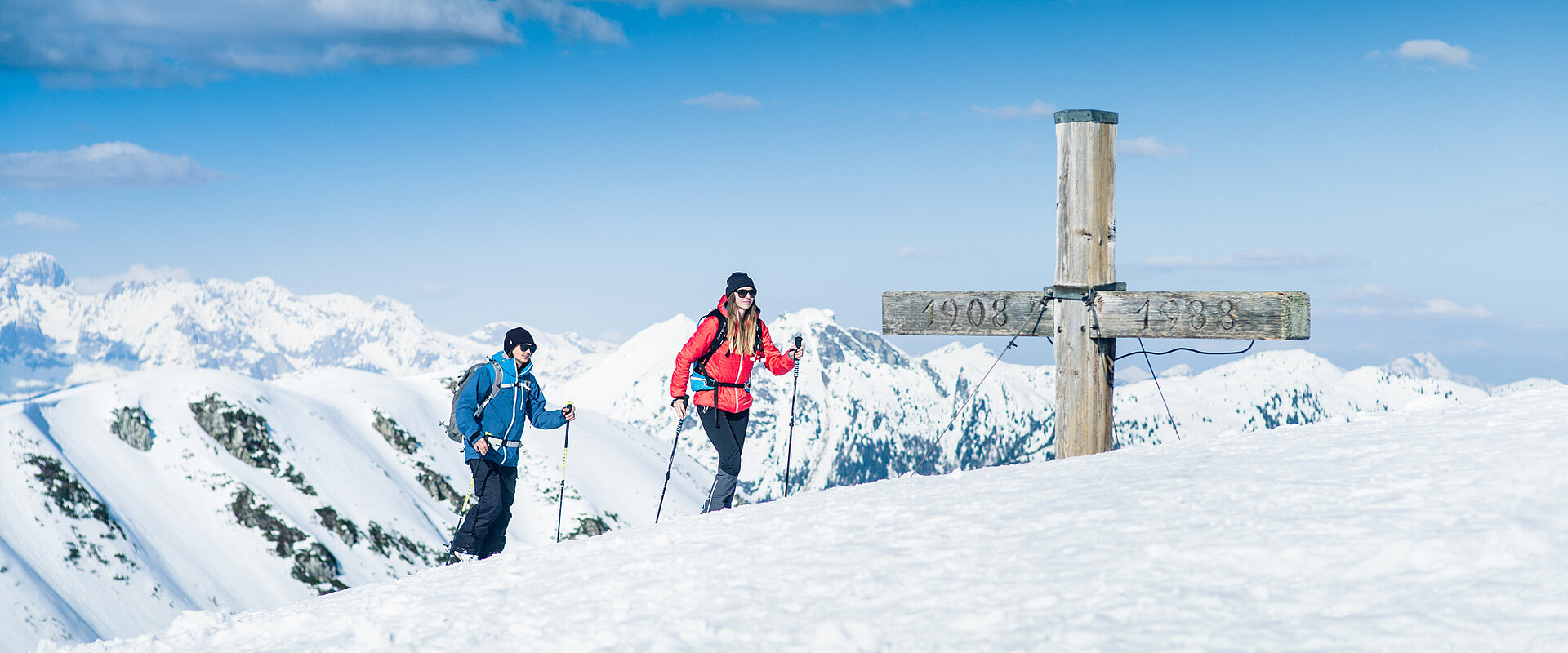 Two ski tourers at the summit cross with panoramic views of the snow-covered Alps in Obertauern