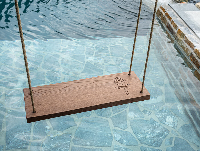 Eine Holzschaukel mit eingraviertem Logo über dem Naturpool im Wellnesshotel Lürzerhof in Salzburg.